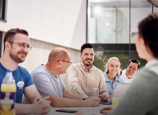 Fünf Personen sitzen an einem Tisch in einem modernen Büro, diskutieren, lächeln und arbeiten gemeinsam an Laptops.
