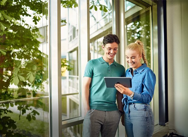 Ein junger Mann und eine junge Frau, stehen in einem Raum vor großen Fenstern, lächeln und schauen auf ein Tablet.