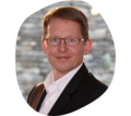 Mann in schwarzem Sakko und weißem Hemd, mit Brille vor unscharfem Hintergrund.
