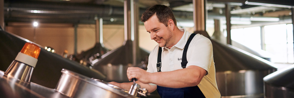 Mann in weißem T-Shirt und blauer Latzhose bedient Ausrüstung in der Krombacher Brauerei mit großen Edelstahltanks.