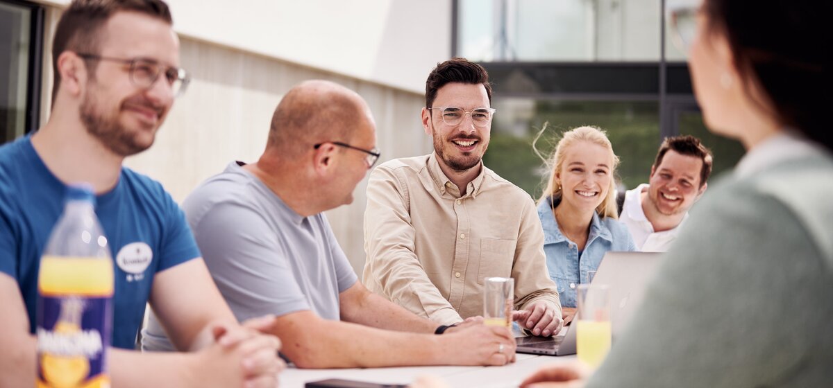 Fünf Personen sitzen an einem Tisch in einem modernen Büro, diskutieren, lächeln und arbeiten gemeinsam an Laptops.