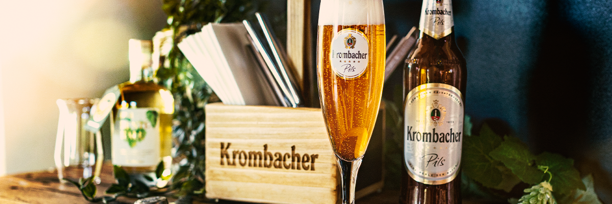 Krombacher Pils Bierflasche und gefülltes Bierglas auf Holztheke neben Holzbox mit Krombacher-Schriftzug und Barutensilien