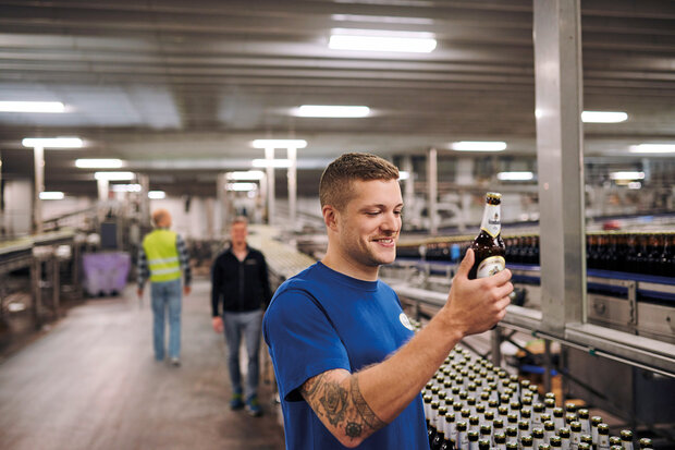 Mitarbeiter in blauer Arbeitskleidung prüft Krombacher Pils Flasche in einer Abfüllanlage mit vielen Flaschen auf dem Förderband.
