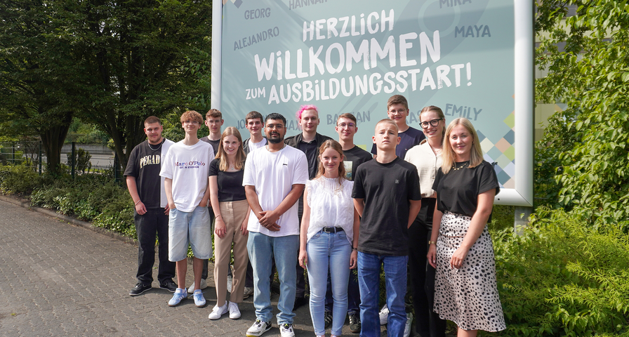 13 junge Menschen stehen vor einem großen Werbeschild auf dem steht 'Herzlich Willkommen zum Ausbildungsstart!'.