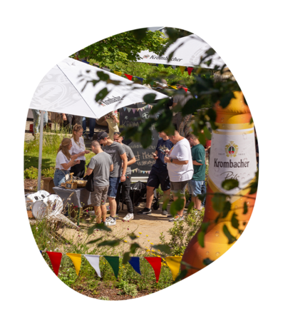 Menschen stehen unter Sonnenschirmen mit Krombacher-Logo beim Krombacher Sommerfest im Garten, bunte Wimpel und Blätter hängen im Vordergrund