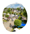Sommerfest mit vielen Menschen, weißen Sonnenschirmen und bunten Wimpeln vor der Krombacher Erlebniswelt, im Hintergrund das Dorf mit Kirche und Wohnhäusern