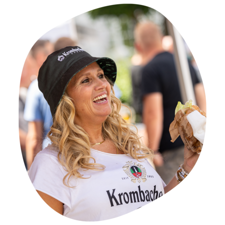 Frau mit langen blonden Haaren trägt Krombacher-Mütze und T-Shirt, hält ein Burger in der Hand